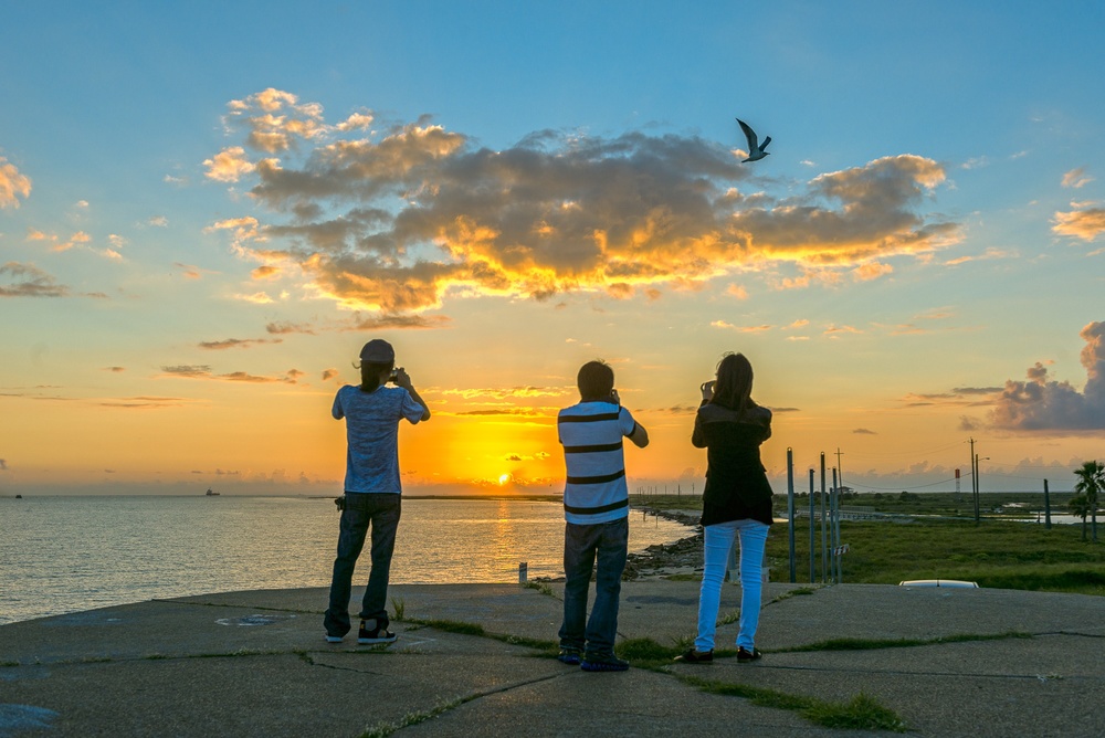 Three people photographing a vibrant sunrise at the East End Lagoon Nature Preserve with a bird flying overhead.