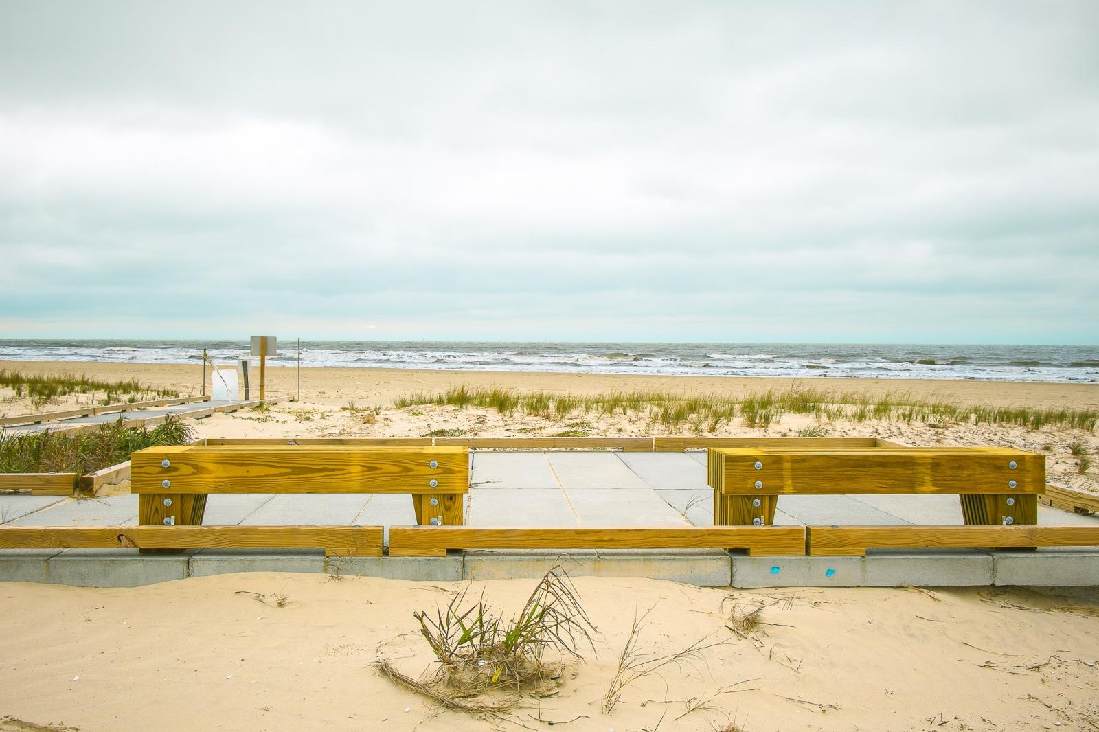 A sandy pathway leading to a calm, open beach with gentle waves at Galveston Island State Park under a cloudy sky.