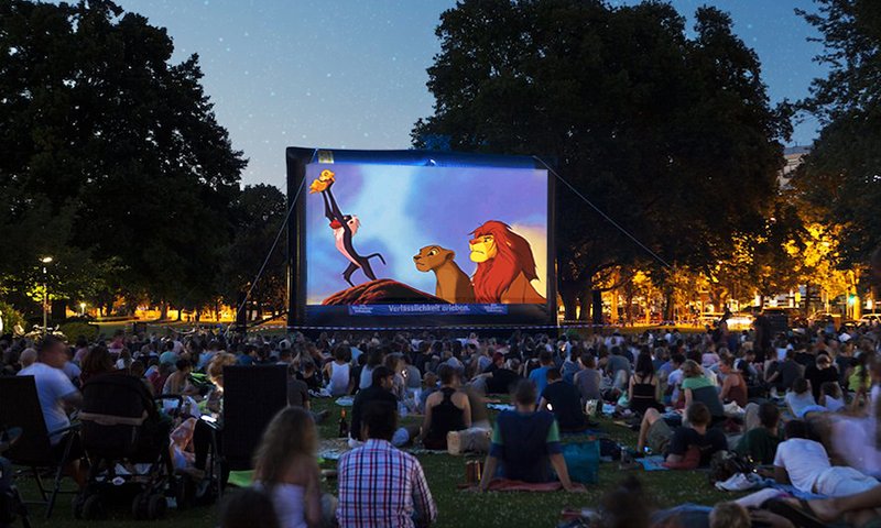 A large group of families and children gathered in a park watching a screening of The Lion King at an outdoor movie night.