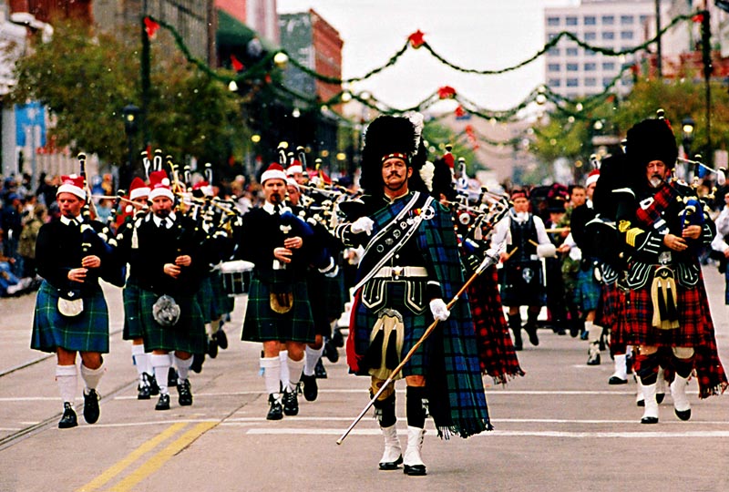 A costumed marching band in traditional Scottish attire parades down a decorated Galveston street during the Dickens on the Strand festival.