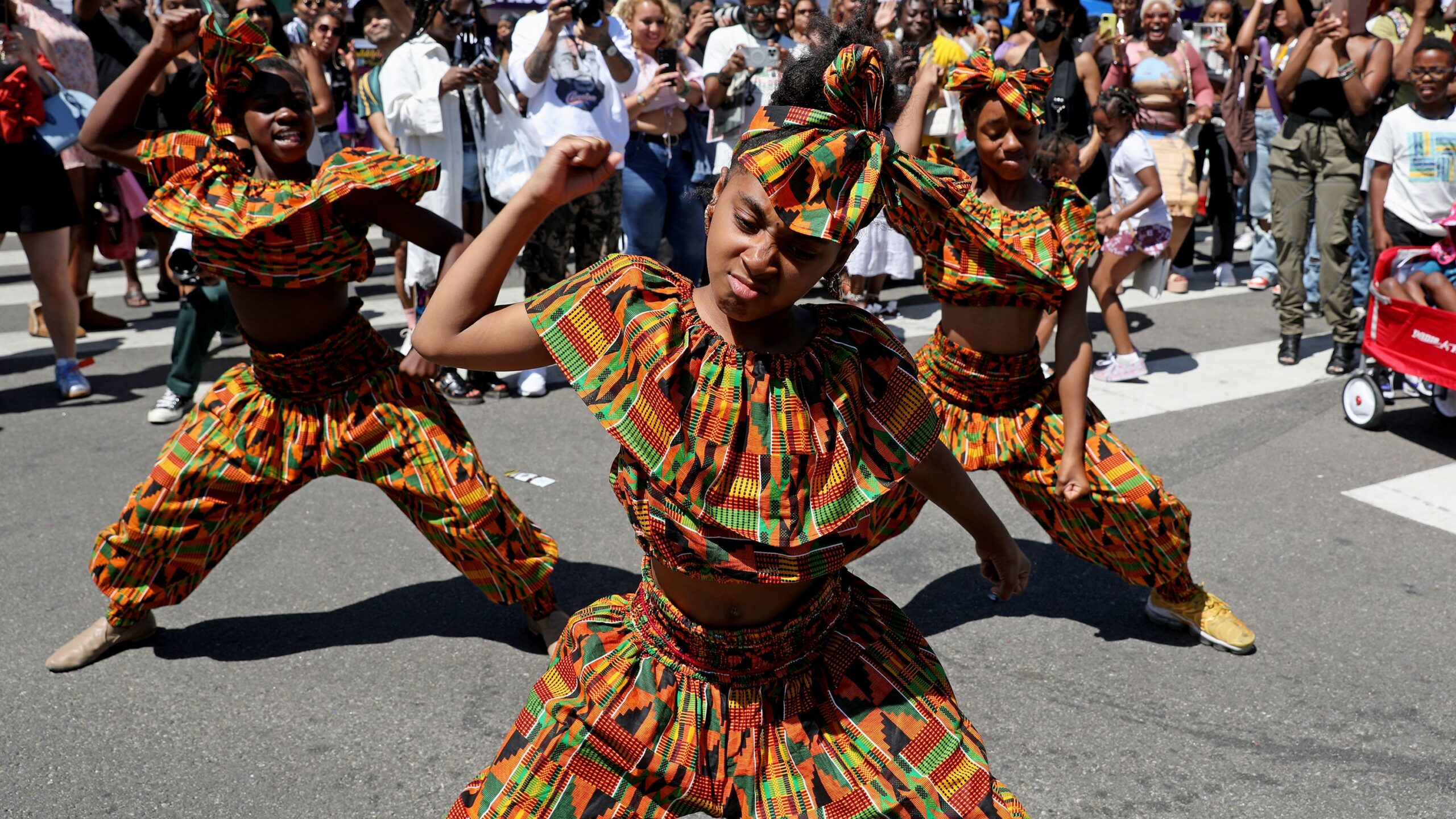 Young performers in colorful African attire dancing energetically during a Juneteenth celebration surrounded by a cheering crowd.