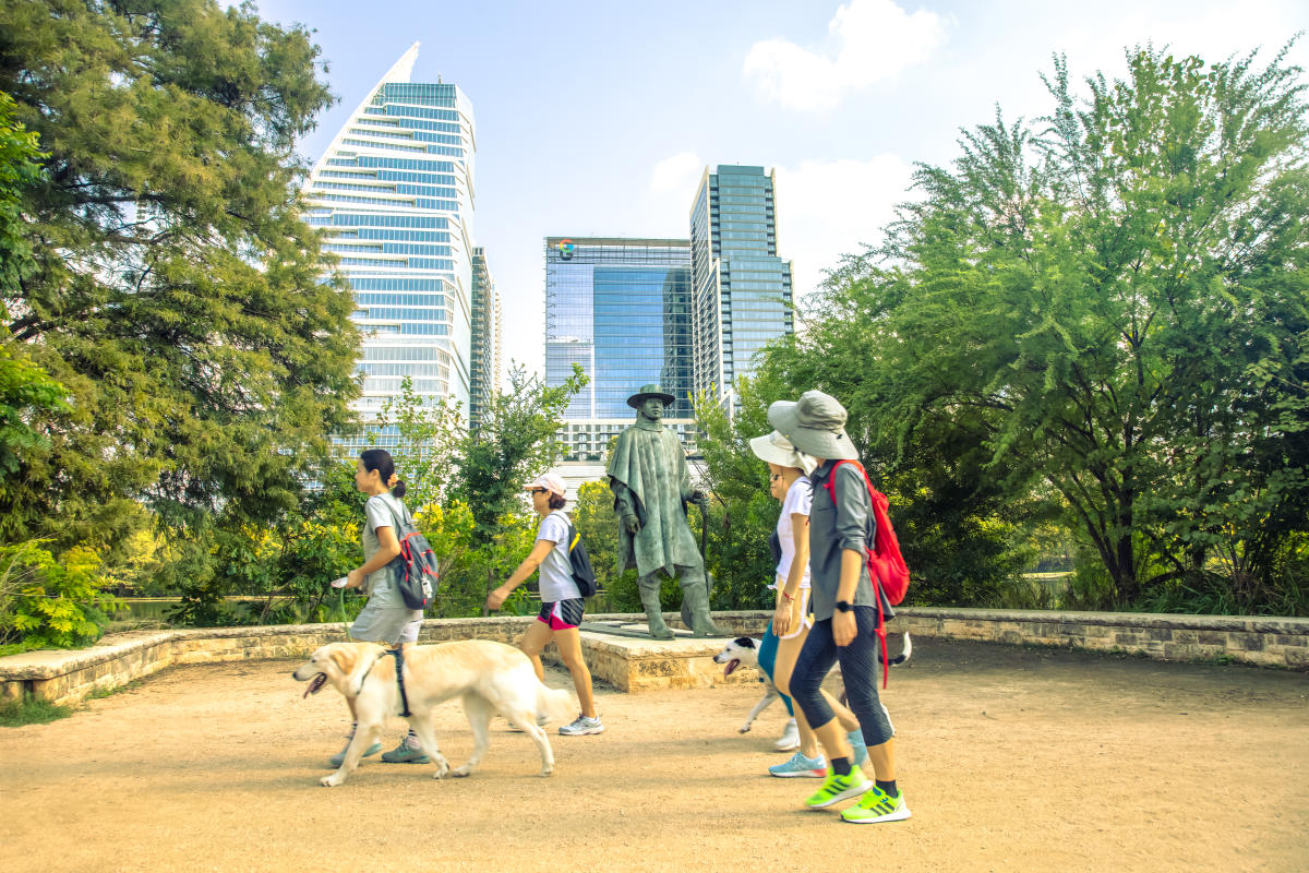 Group of people walking dogs near the Stevie Ray Vaughan statue with downtown Austin skyline in the background at Lady Bird Lake Trail.