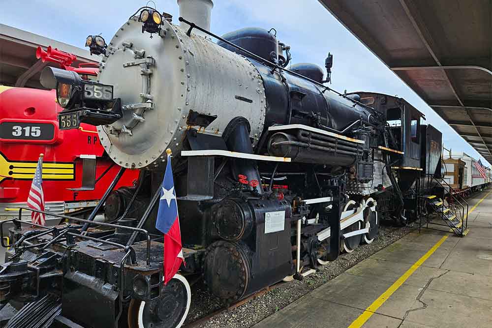 A historic black steam locomotive and a bright red train displayed at the Galveston Railroad Museum’s platform.
