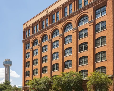 Exterior view of the red-brick Sixth Floor Museum at Dealey Plaza with the Reunion Tower visible in the background against a blue sky.