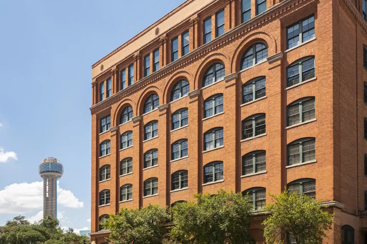 Exterior view of the red-brick Sixth Floor Museum at Dealey Plaza with the Reunion Tower visible in the background against a blue sky.