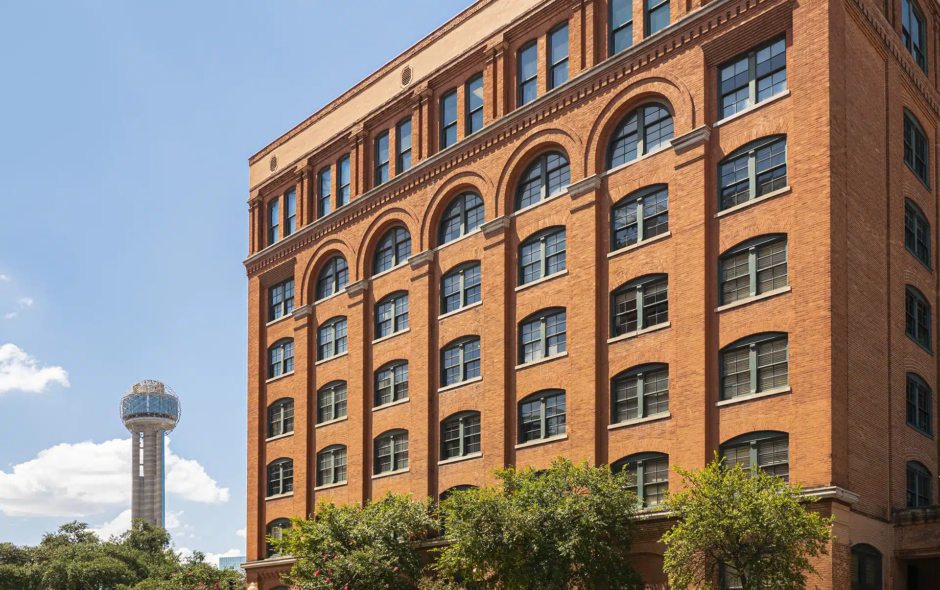 Exterior view of the red-brick Sixth Floor Museum at Dealey Plaza with the Reunion Tower visible in the background against a blue sky.