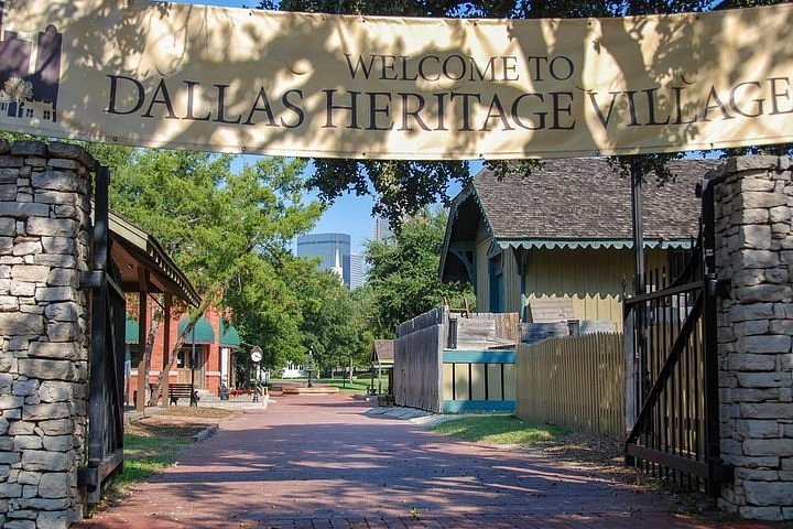 Entrance to Dallas Heritage Village featuring historic buildings and a tree-lined brick path under a welcome banner.