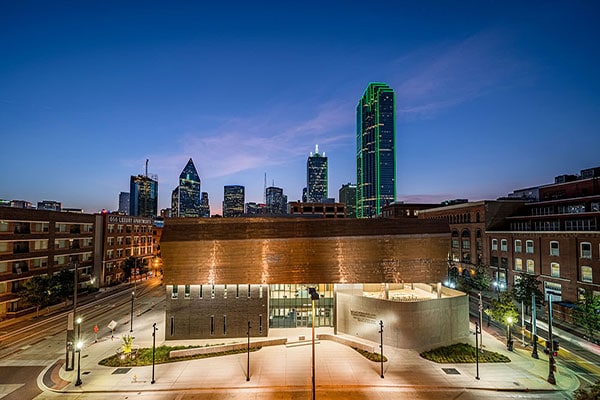 Exterior view of the Dallas Holocaust and Human Rights Museum at dusk with the Dallas skyline in the background.