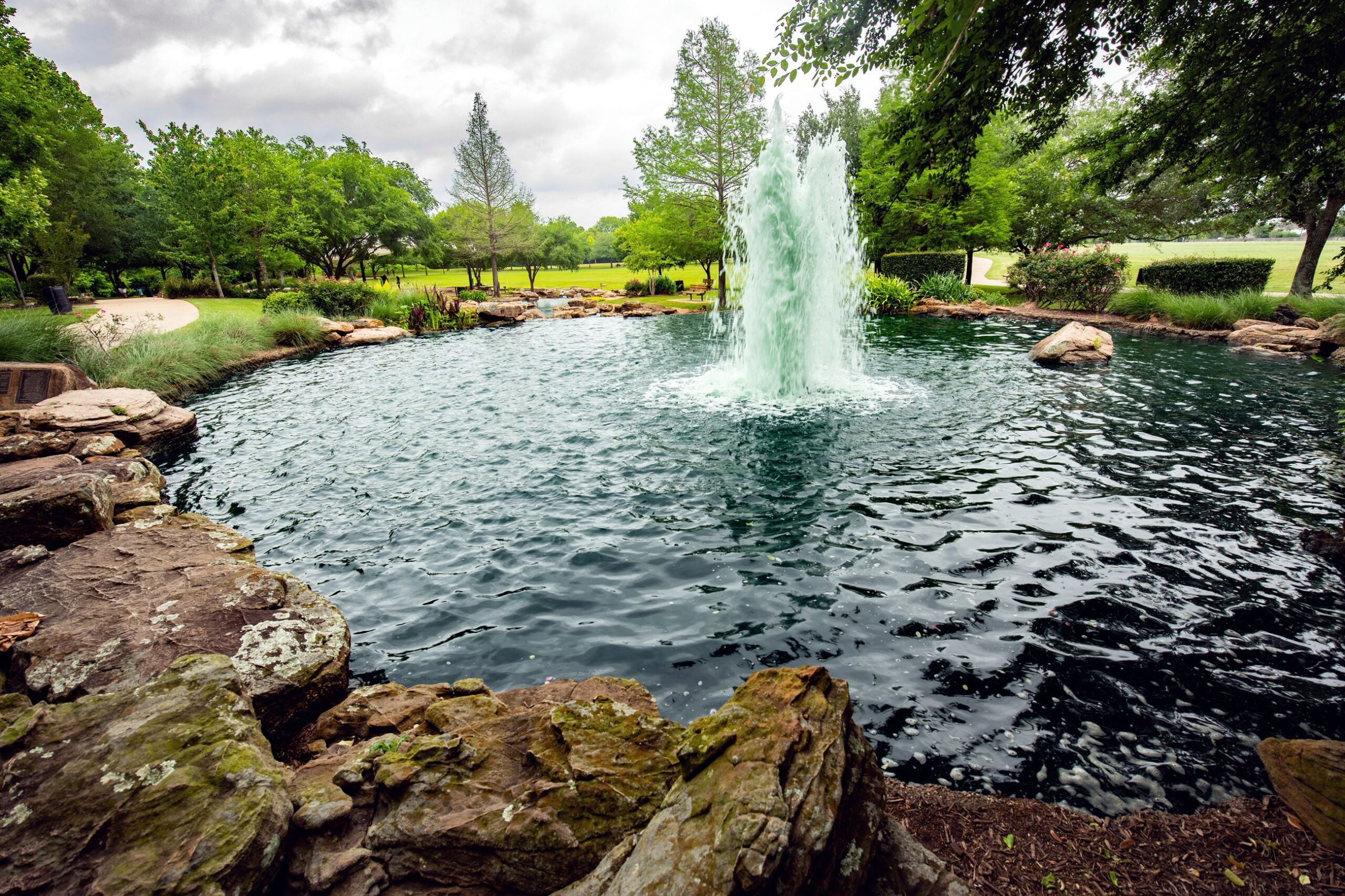 Scenic view of a fountain in the middle of a pond surrounded by trees and stone walkways at Oyster Creek Park in Sugar Land.