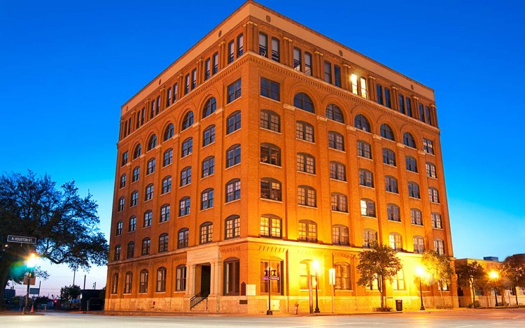 The red-brick Sixth Floor Museum building at Dealey Plaza in Dallas, illuminated at twilight.