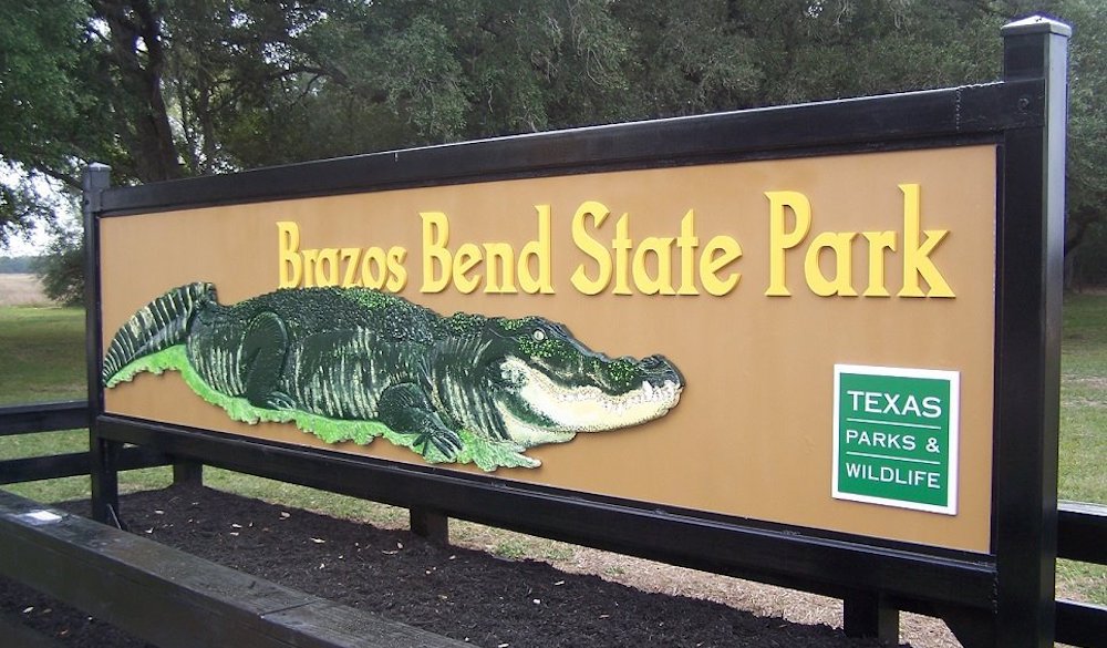 Brazos Bend State Park entrance sign featuring an alligator illustration, surrounded by trees.