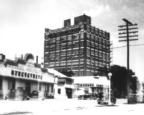 Black and white photo of early Sugar Land featuring the Imperial Sugar refinery, vintage vehicles, utility poles, and storefronts.