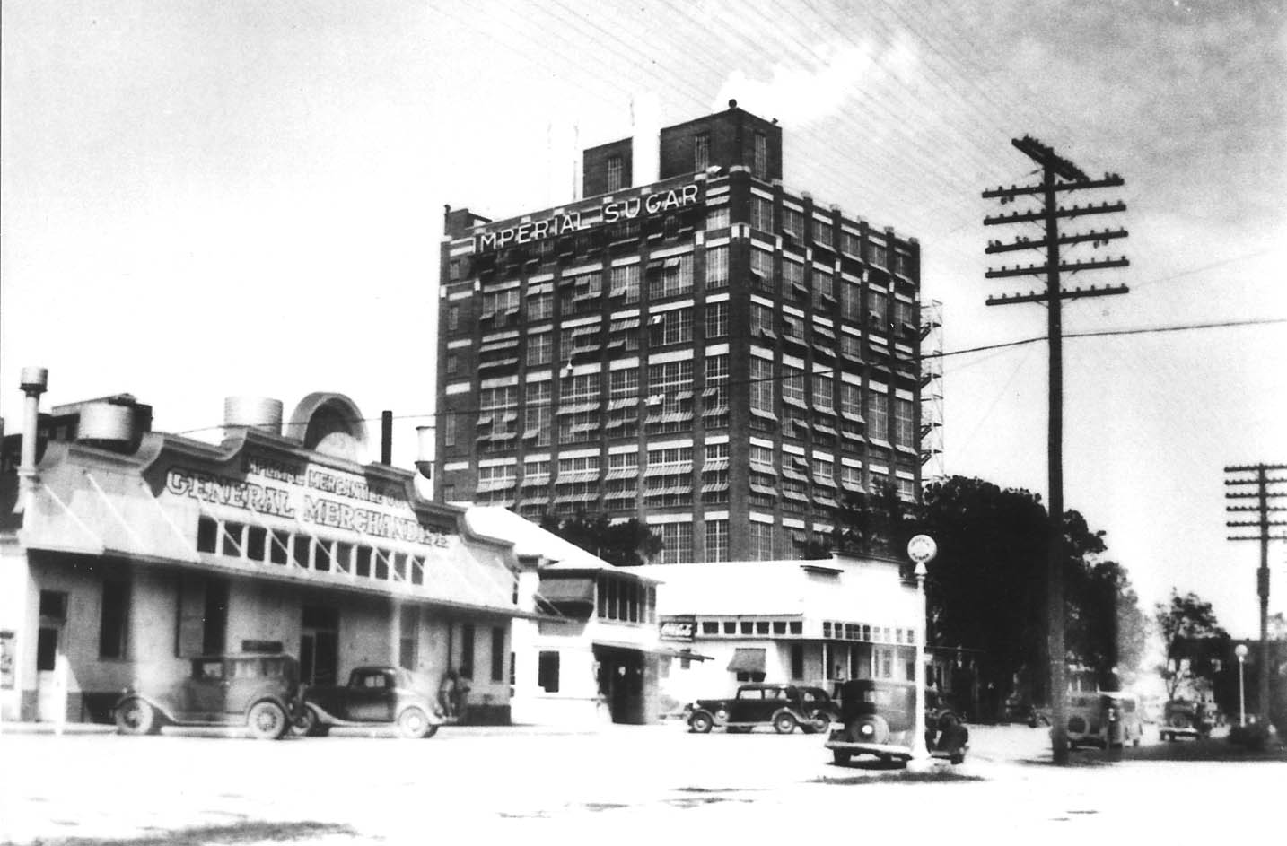 Black and white photo of early Sugar Land featuring the Imperial Sugar refinery, vintage vehicles, utility poles, and storefronts.