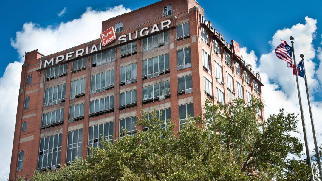 Modern view of Imperial Sugar Refinery building with American and Texas flags waving in the foreground under a blue sky.