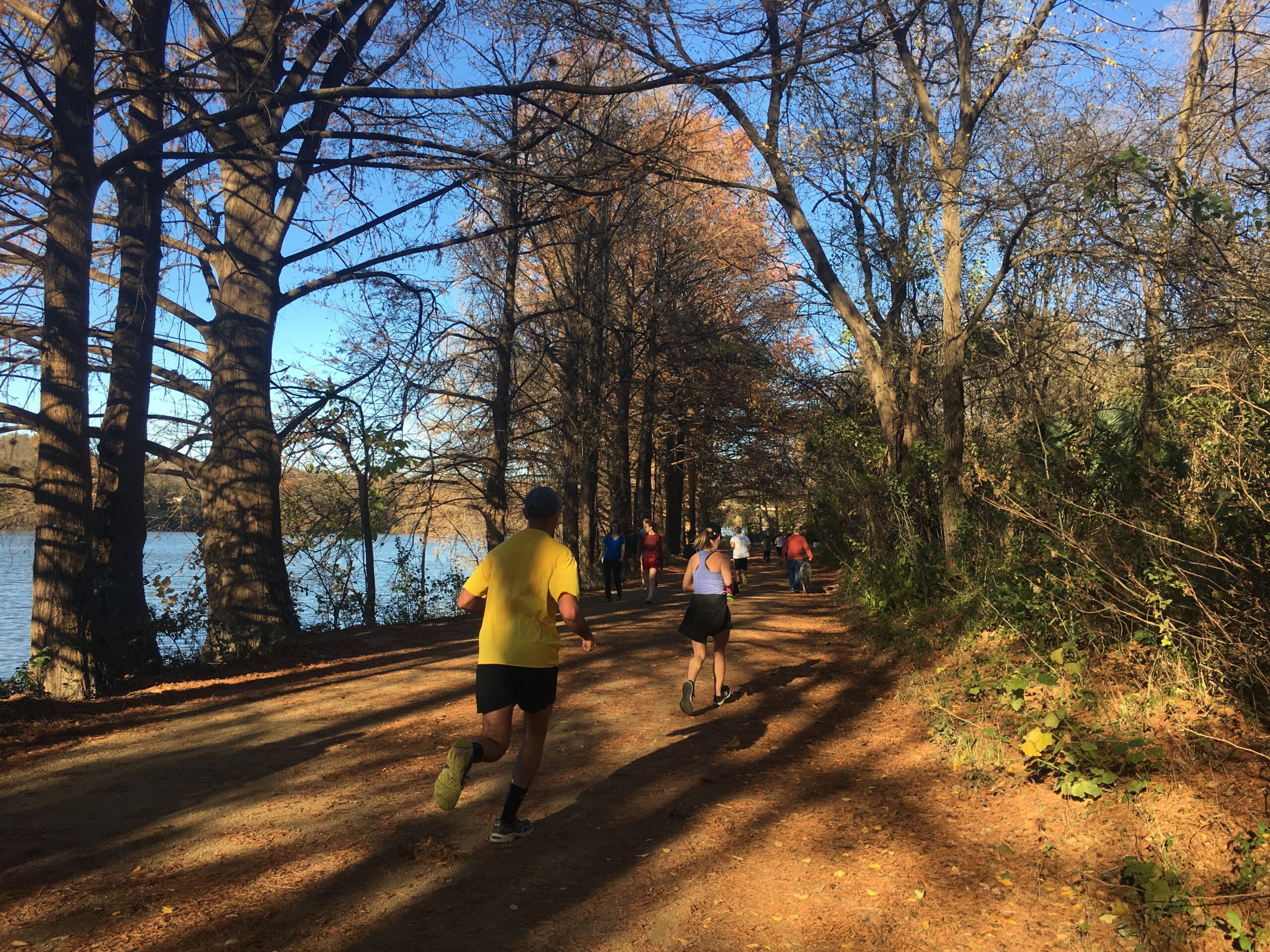 People jogging and biking along the shaded trail at Lady Bird Lake in Austin with trees and water in the background.