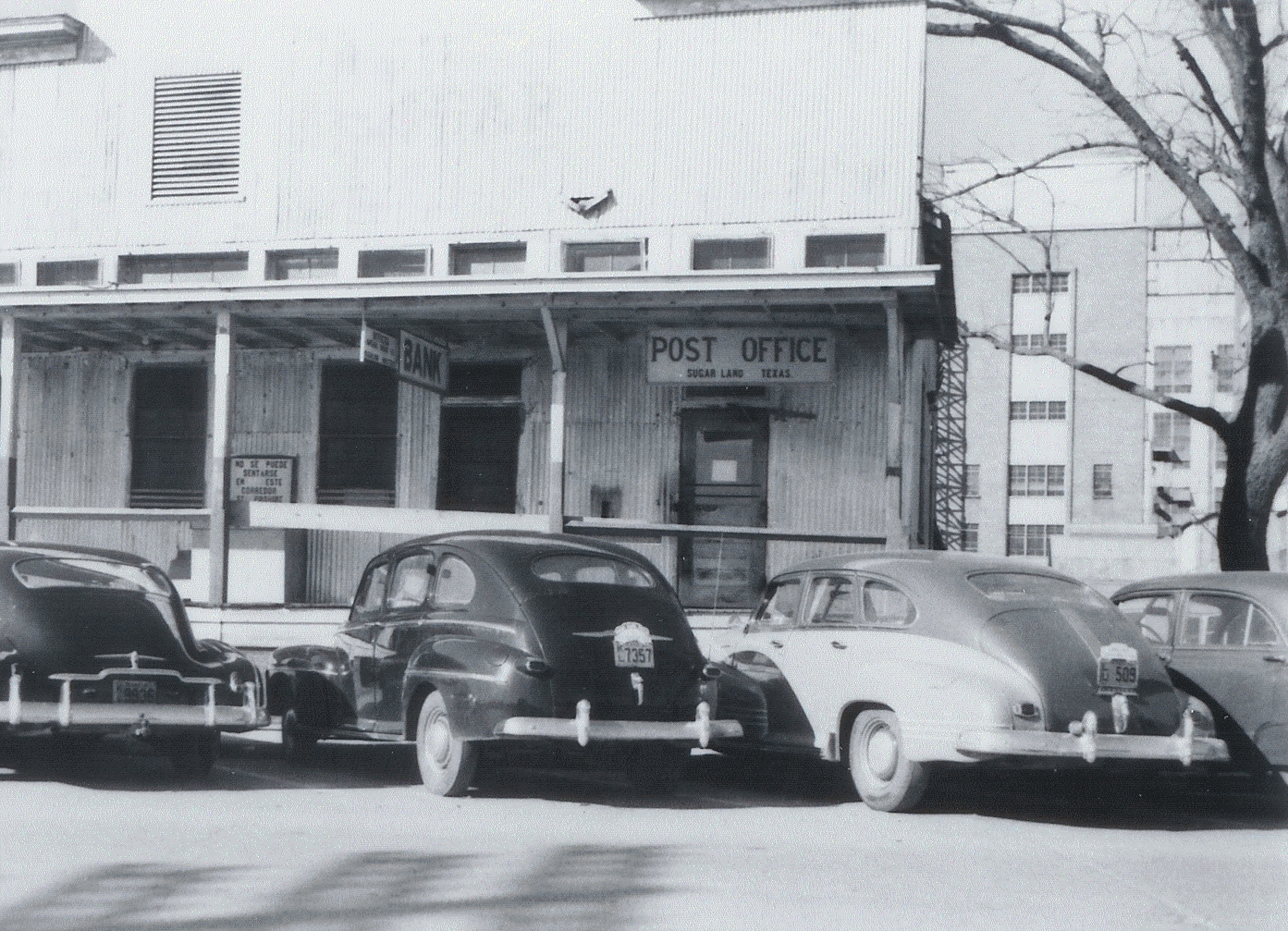 Historic photo of Sugar Land, Texas post office and bank building with vintage cars parked in front, circa 1940s.