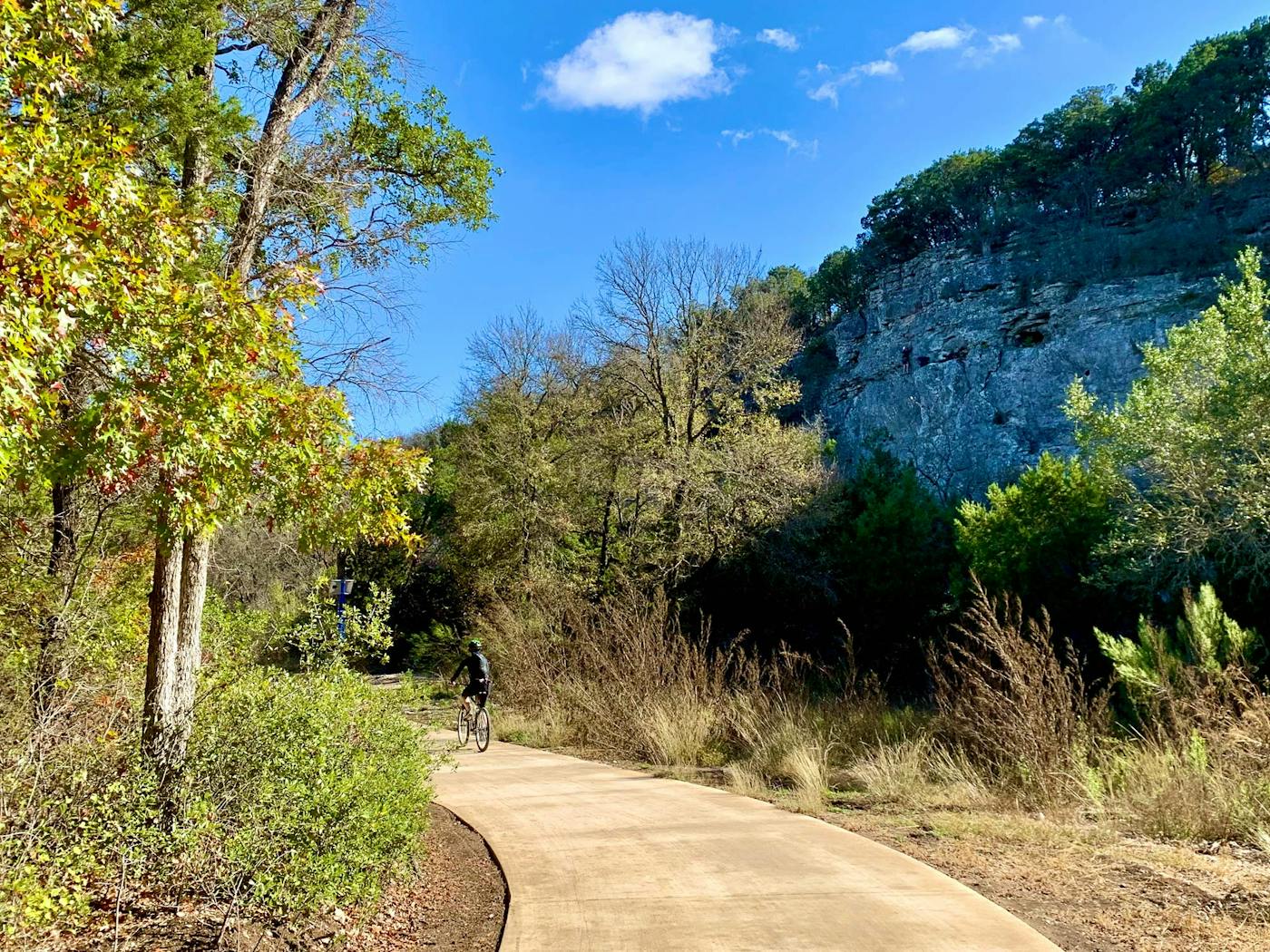 Biker on a paved trail next to rocky cliffs and trees along the San Antonio Mission Trail in Texas.