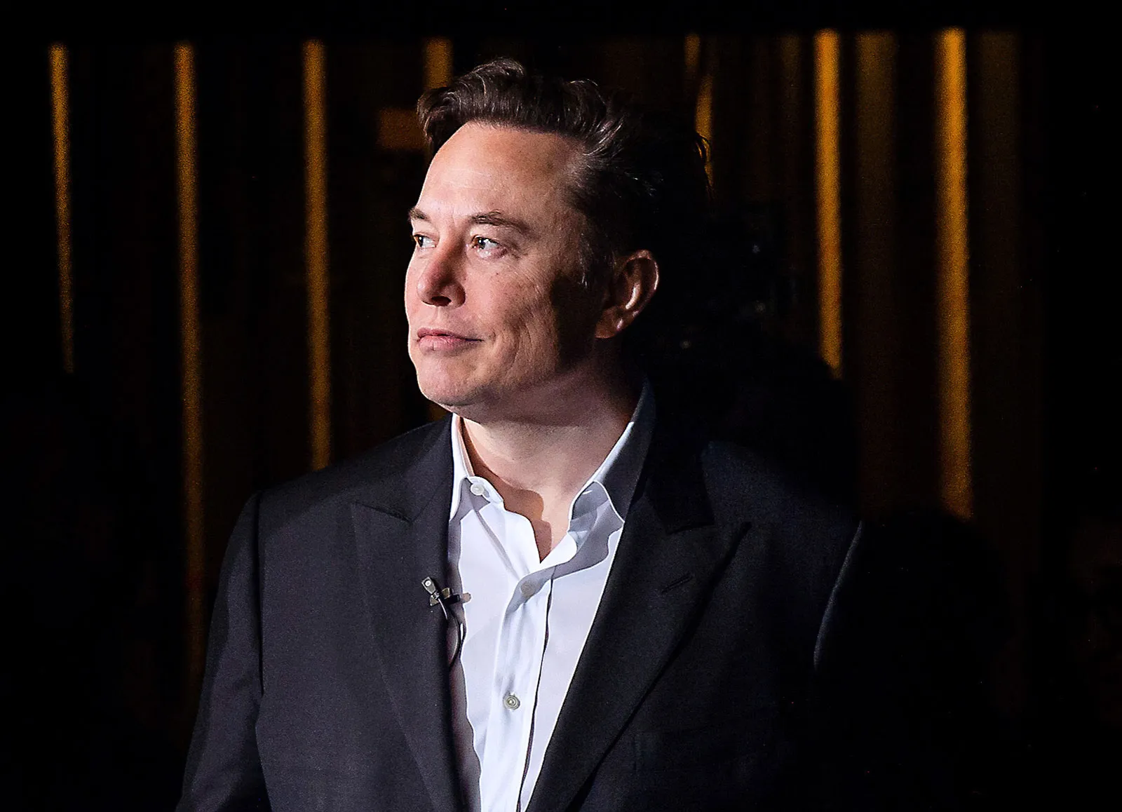 Elon Musk wearing a suit, looking to the side under dramatic lighting in Texas.